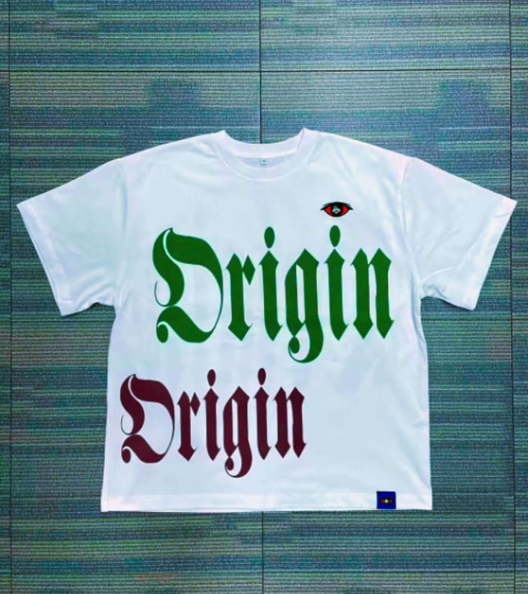 IBeorigin T-Shirt (Pre-Order) featuring bold 'Origin' text in green and red gothic font with an eye graphic on white cotton fabric.
